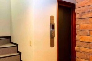 L’entrée d’une chambre d’hôtel, avec une porte en bois, un bouton d’ascenseur fixé au mur et un escalier menant à un étage supérieur.