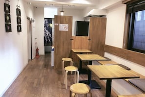 A restaurant interior with wooden tables and stools, a counter, and a window with a view. - Guest House Siesta (Tokyo)