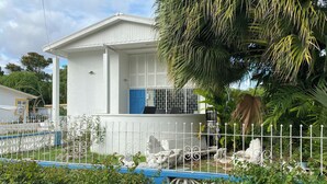 A white building with a blue door, surrounded by a white fence and lush greenery. - 3-bedroom Coastal Escape in lovely Christ Church with Wi-Fi, AC (Bridgetown)
