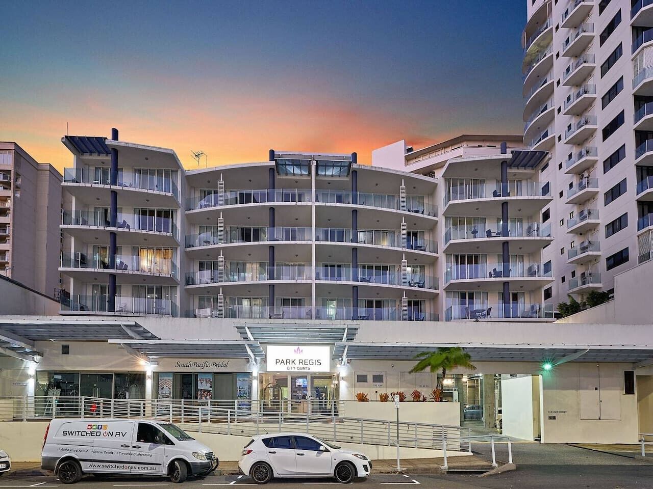 A multi-story hotel with a modern design, featuring balconies on each floor, a covered entrance, and a sign for 'Park Regis City Quays.'