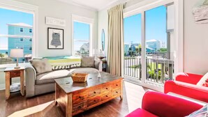 A spacious living room with a wooden coffee table, a grey sofa, and a red armchair. Large windows offer a view of the city and a balcony with a railing.