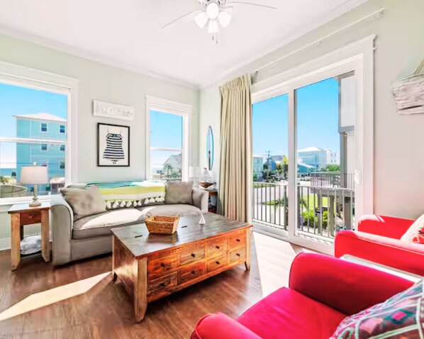 A spacious living room with a wooden coffee table, a grey sofa, and a red armchair. Large windows offer a view of the city and a balcony with a railing.