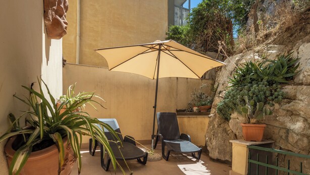 Een zonnig buitengebied met zitplaatsen, een tafel, stoelen, een grote parasol en potplanten.