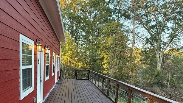 A wooden deck with a railing, a red building with windows, and trees in the background. - Wandering Inn FY25 Build. (Monroe)