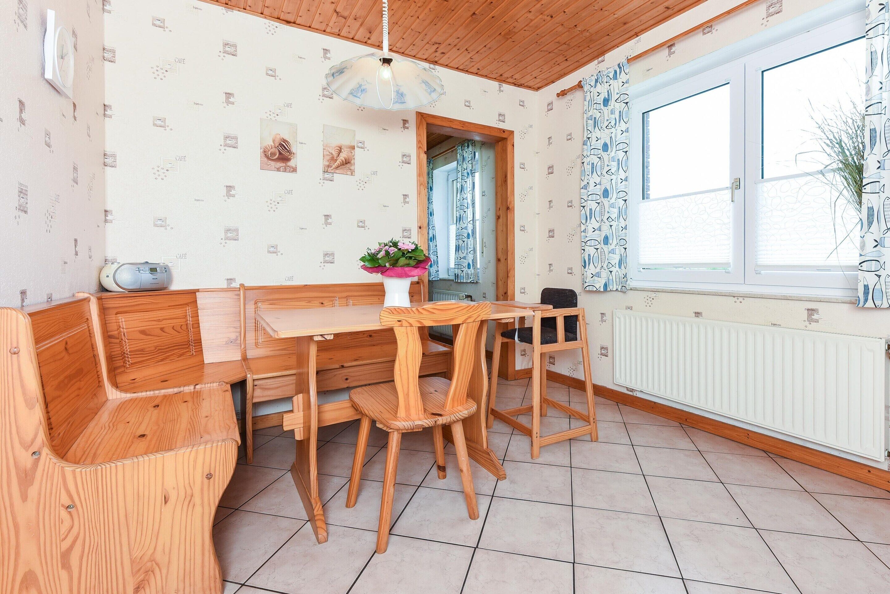 A wooden dining area with a bench, a table, and chairs. There is a radiator on the wall and a window with curtains.