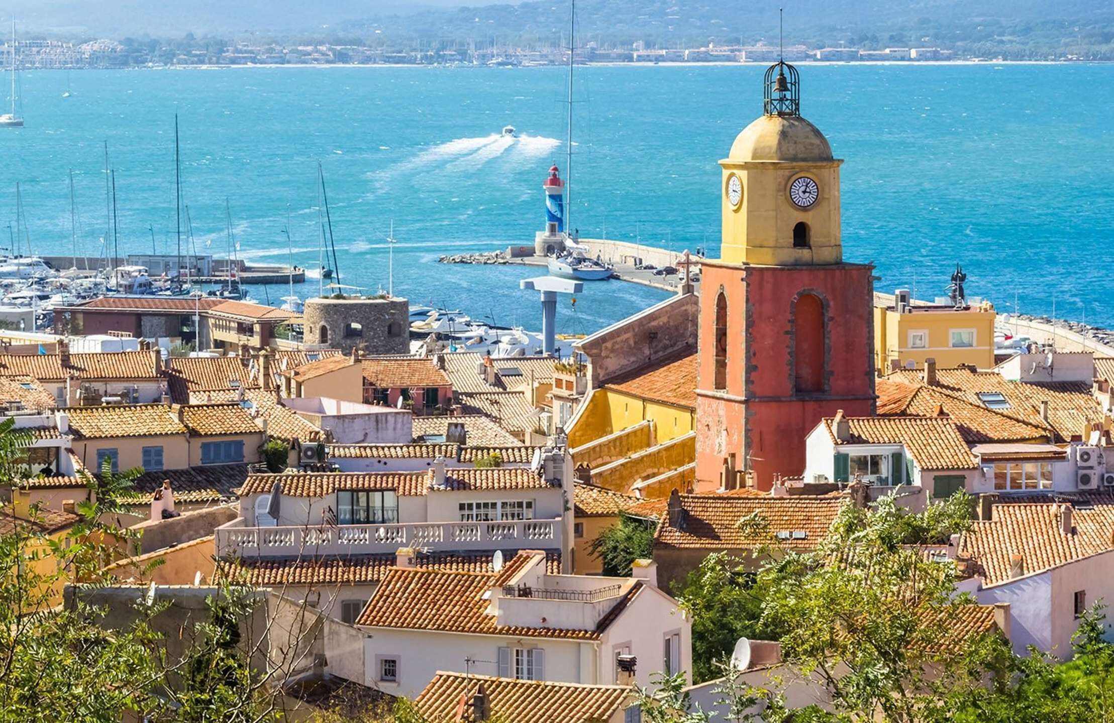 A coastal town with a prominent clock tower, a marina with boats, and terracotta-roofed buildings.