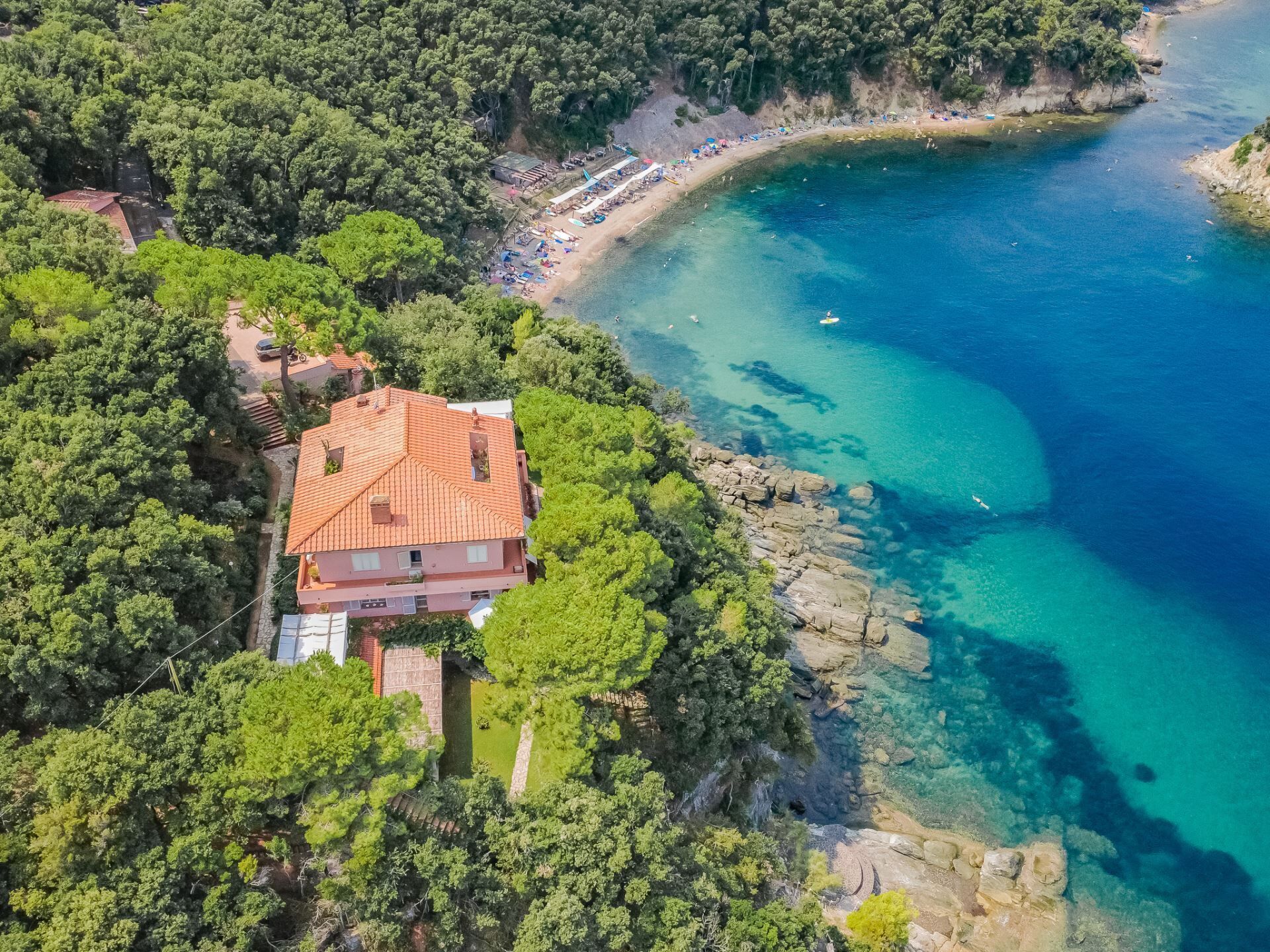 Una proprietà costiera con una casa a tetto rosso immersa nel verde, affacciata su un mare azzurro e una spiaggia sabbiosa.