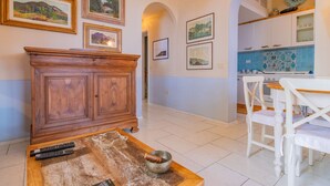 Ein Wohnzimmer mit Holzschrank, Couchtisch mit Schale und Fernbedienungen sowie eine Küchenzeile mit weißen Schränken und blauen Fliesen.
