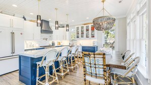 A modern kitchen with white cabinets, a large island, and a dining area with wicker chairs. - Coastal Retreat w/ Private Pool | Golf Cart (Oak Island)