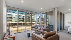 A modern living room with a large window, a sofa set, a coffee table, and a flat-screen TV. - Spacious Lakefront Apartment With Pool Access (Canberra)