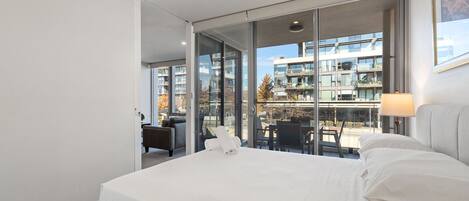 A modern hotel room with a large bed, a glass wall offering a view of the cityscape, and a small balcony with seating.