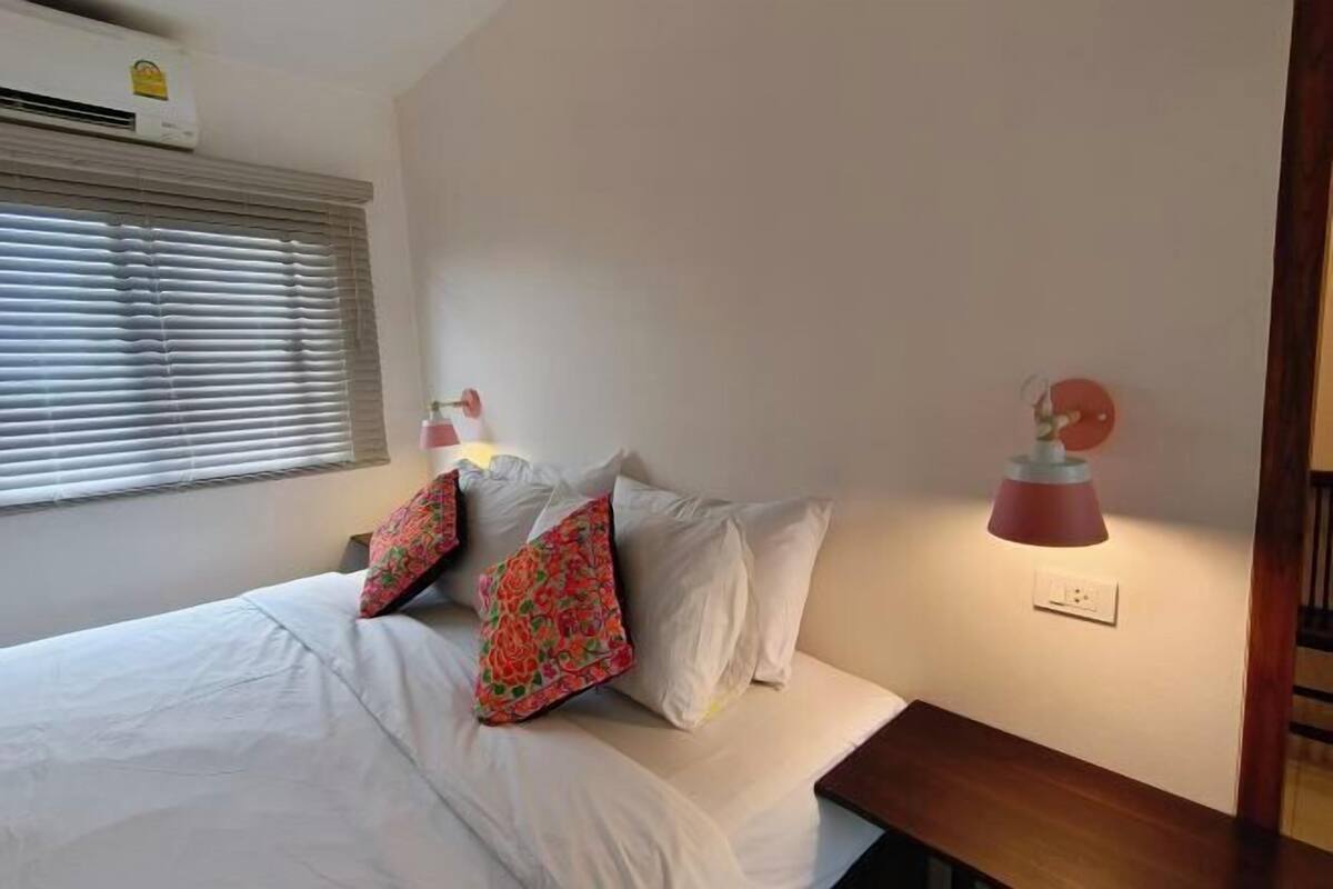A neatly made bed with white linens and decorative pillows, a bedside table with a lamp, and a window with blinds.