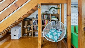 Una camera mansardata accogliente, con travi a vista, scaffali di legno, una poltrona bianca con cuscino decorato e un divano verde.