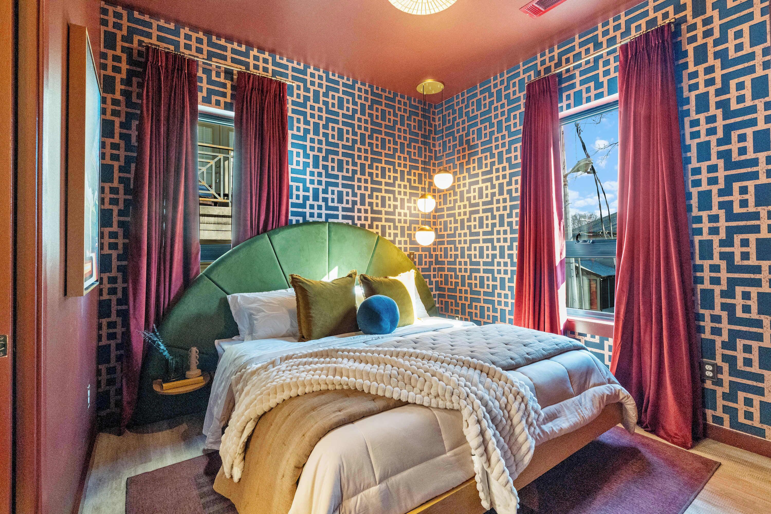 A bedroom with a green upholstered headboard, a large bed with multiple pillows, a patterned wall, and a window with red curtains.