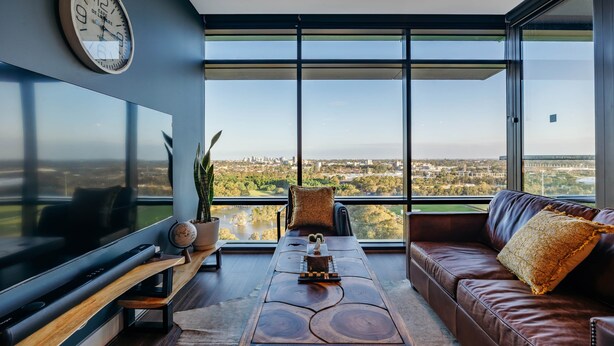 A modern living room with a large TV, a wooden coffee table, a brown leather sofa, and a view of a cityscape through large windows.