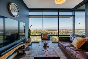 A modern living room with a large TV, a wooden coffee table, a brown leather sofa, and a view of a cityscape through large windows. - "Horizon" King-bed Lux Apt Stunning View w Parking (Sydney Olympic Park)