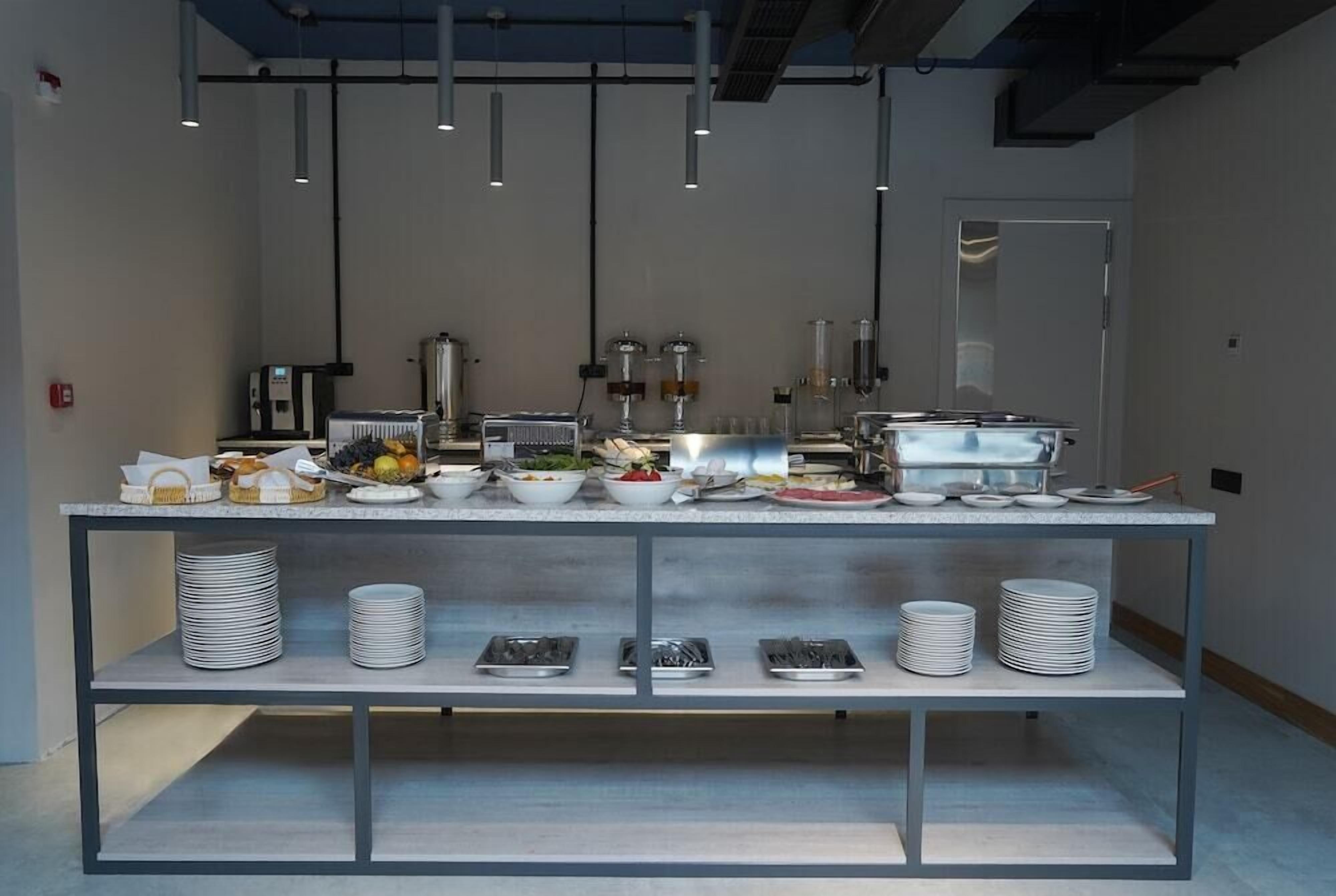 Disposición de un bufé con diferentes alimentos y utensilios sobre una mesa larga, con iluminación cenital y una puerta al fondo.