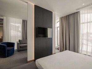 A modern hotel room with a bed, a TV mounted on a wall, a seating area with a chair, and large windows with curtains. - Radisson Hotel & Spa Gyumri (Gyumri)
