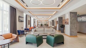 A modern hotel lobby with a central seating area, a reception desk, and a large window providing natural light.