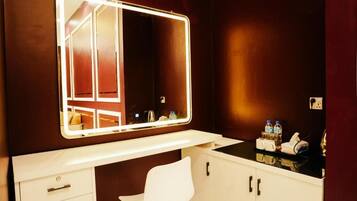 A bathroom with a vanity, a mirror with lights, a white chair, and toiletries on the counter.
