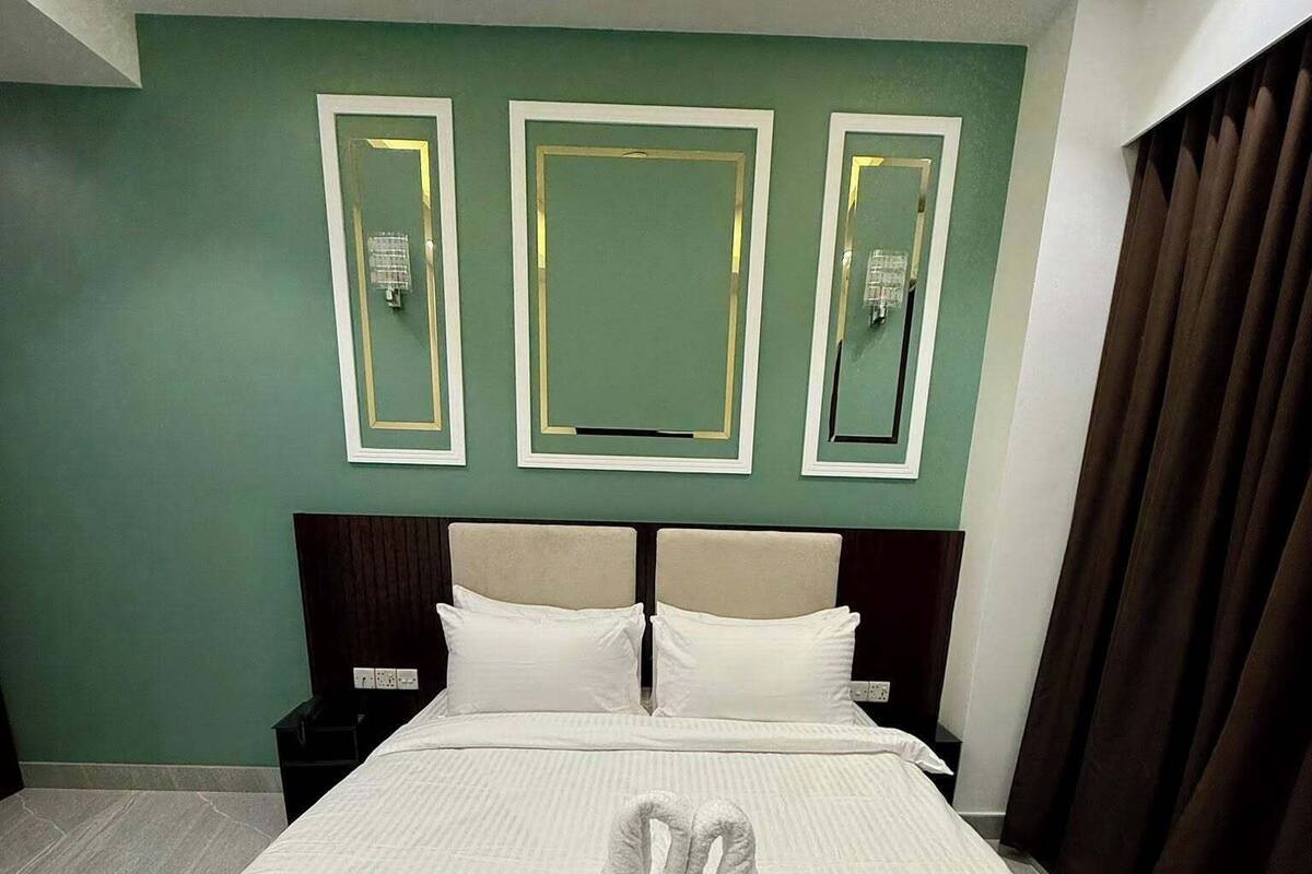 A neatly made bed with a towel swan centerpiece, a dark headboard, and a green wall with three framed mirrors.