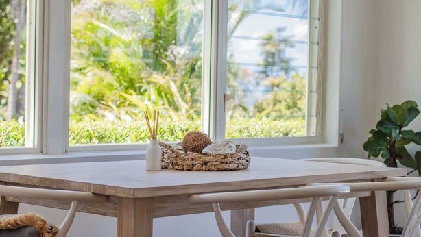 Een houten tafel met een geweven mand en kandelaar, geplaatst nabij een raam met uitzicht op groen.