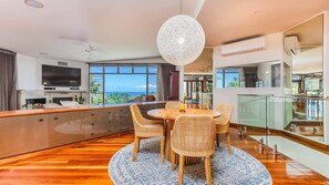 Un salon moderne avec une table à manger ronde, des chaises en osier, un grand luminaire suspendu et une vue sur l’océan.