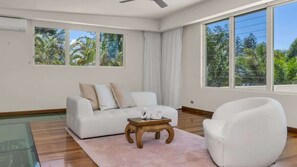 Ein modernes Wohnzimmer mit einem weißen Sofa, einem Holztisch und einem weißen Sessel. Große Fenster bieten Blick auf viel Grün.