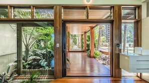 Un intérieur moderne avec un plancher en bois menant à une porte en verre, un meuble blanc et une vue sur un jardin tropical.
