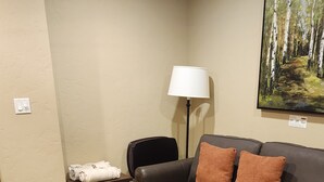 Ett vardagsrum med ett soffbord i trä, en brun lädersoffa med orange kuddar, en svart fåtölj, en golvlampa och en tavla med ett skogsmotiv på väggen.
