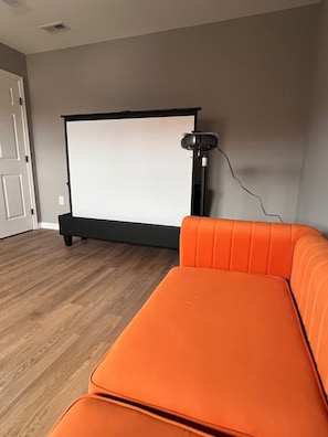 Een kamer met een groot projectiescherm, een zwarte standaard en een oranje bank.