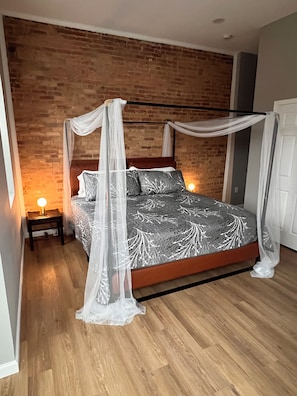 Een bed met hemel, een nachtkastje met lamp, en een bakstenen muur.