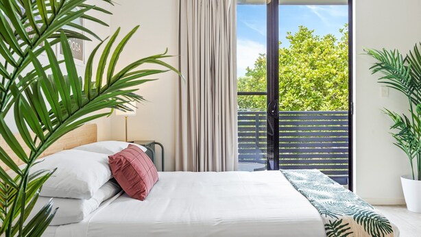 A bedroom with a bed, a window with curtains, a view of greenery outside, and a potted plant. - Comfy Studio Apartment in Marcoola Beach (Sunshine Coast)