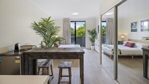 A modern bedroom with a large bed, a small desk, and a dining table. There are potted plants and a sliding glass door to the balcony. - Comfy Studio Apartment in Marcoola Beach (Sunshine Coast)