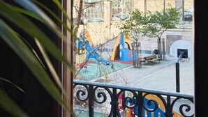 Blick von einem Balkon mit Metallgeländer auf einen Spielplatz mit bunten Spielgeräten und ein Gebäude mit mehreren Fenstern im Hintergrund.