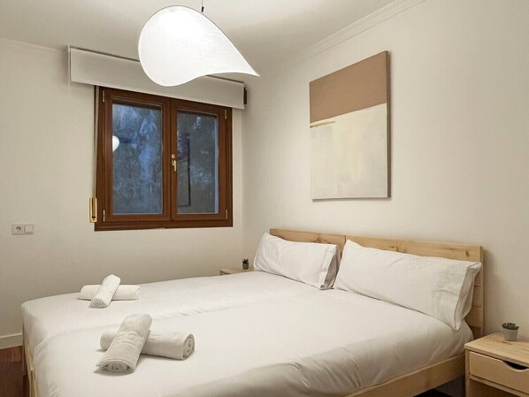 A neatly made bed with white linens and two rolled towels, a window with a view of trees, a ceiling fan, and a small bedside table with a plant.