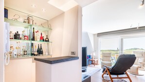 Zona bar moderna con un ripiano in vetro che espone diverse bottiglie, un bancone e un'area salotto con una poltrona e un divano.