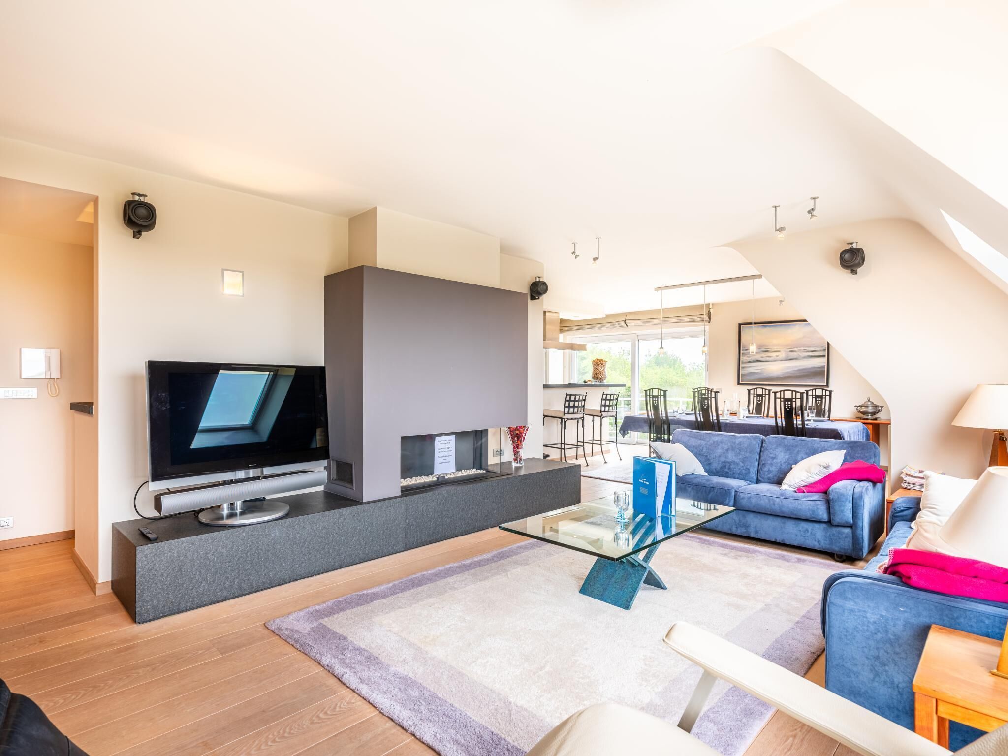 A modern living room with a flat-screen TV, a glass coffee table, a blue sofa, and a dining area with a table and chairs.