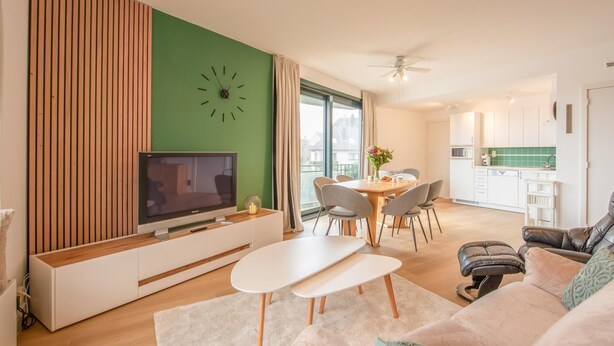 Un salon moderne avec un téléviseur à écran plat, une table à manger avec des chaises, un canapé et une cuisine en arrière-plan.