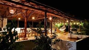 Un patio techado al aire libre con muebles de madera, luces colgantes y una piscina.
