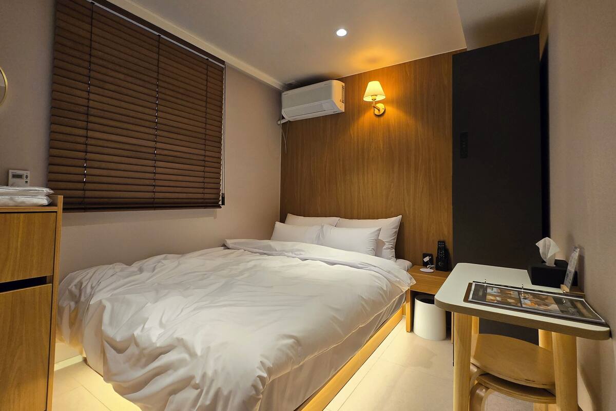 A hotel room with a bed, bedside table, lamp, and a wall-mounted air conditioner.