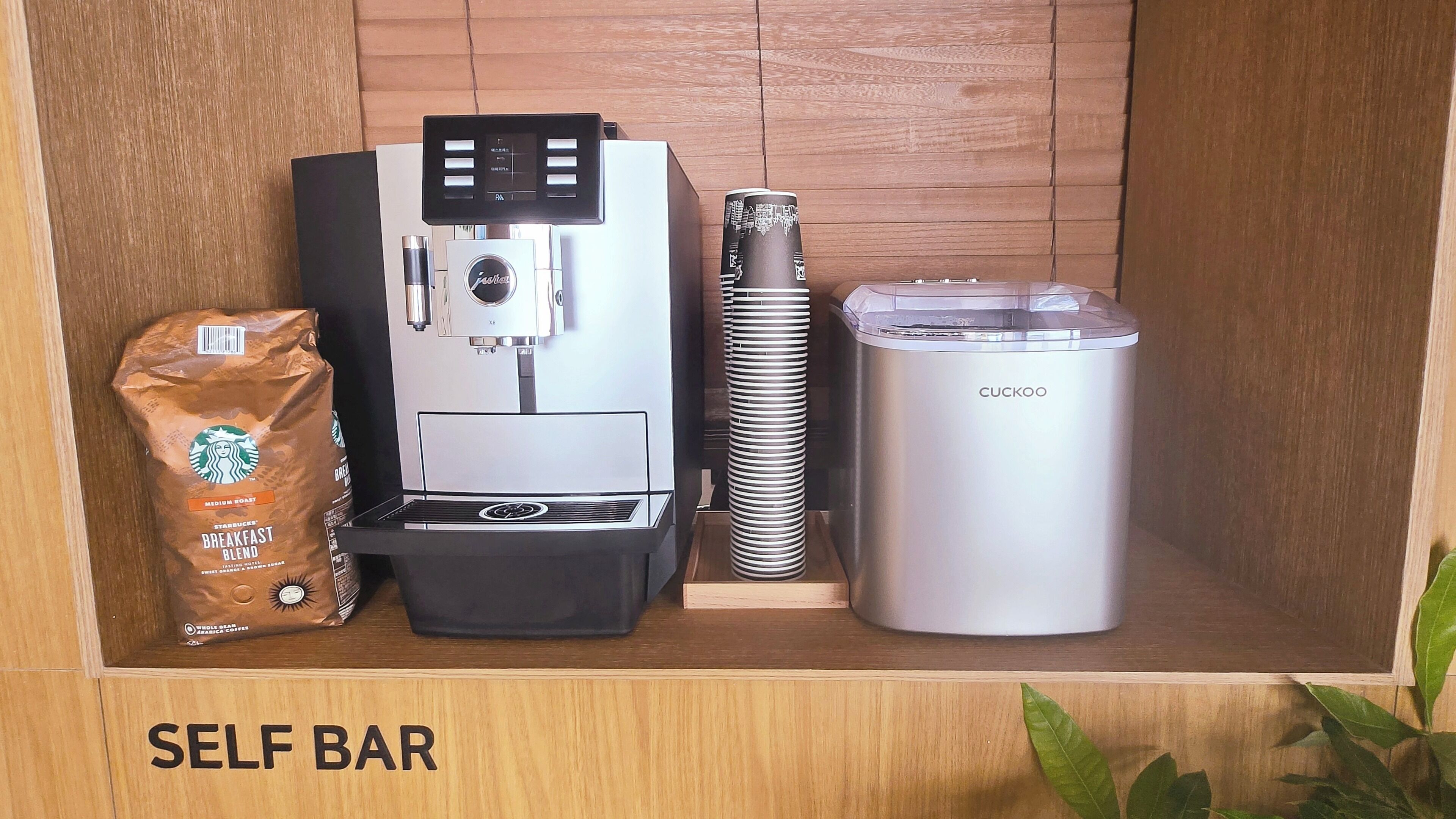 A self-service coffee station with a coffee machine, a coffee pod dispenser, a reusable cup, and a bag of coffee beans.