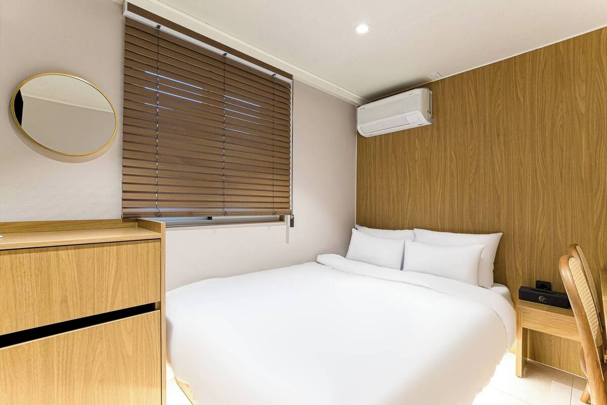 A modern hotel room with a large bed, wooden paneling, a round mirror, and a window with blinds.