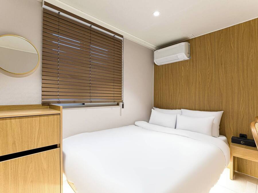 Una habitación de hotel moderna con una cama grande, paneles de madera, un espejo redondo y una ventana con persianas.