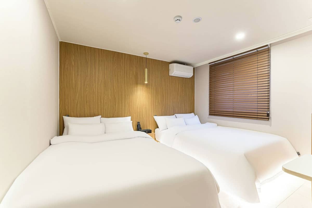 A modern hotel room with two beds, a wooden accent wall, a ceiling-mounted air conditioner, and a window with blinds.