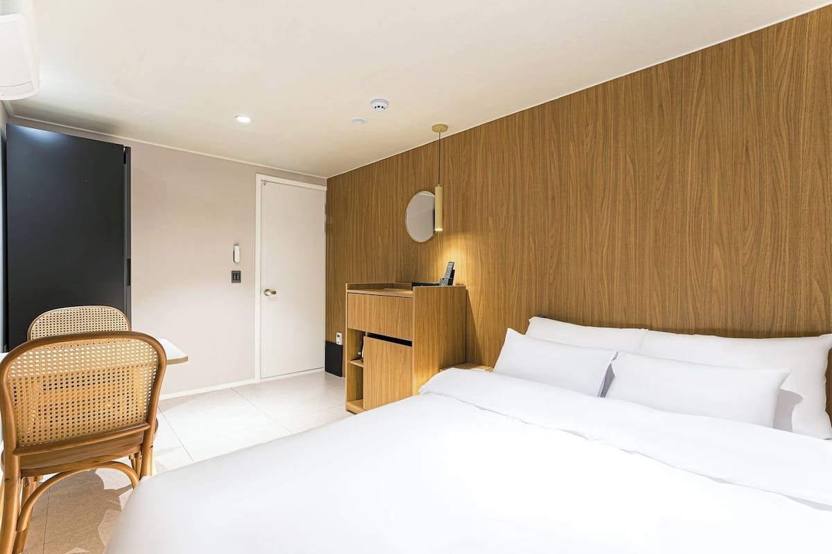 A modern hotel room with a wooden accent wall, a bed with white linens, a wooden nightstand with a mirror, and a wicker chair.