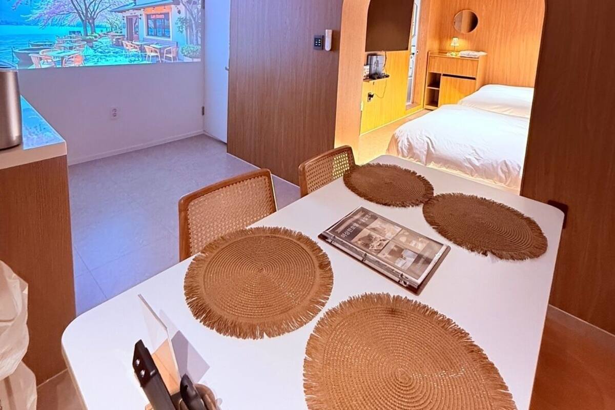A modern hotel room with a dining area, a bed with a wooden headboard, and a large window with a view of a beach and a restaurant.