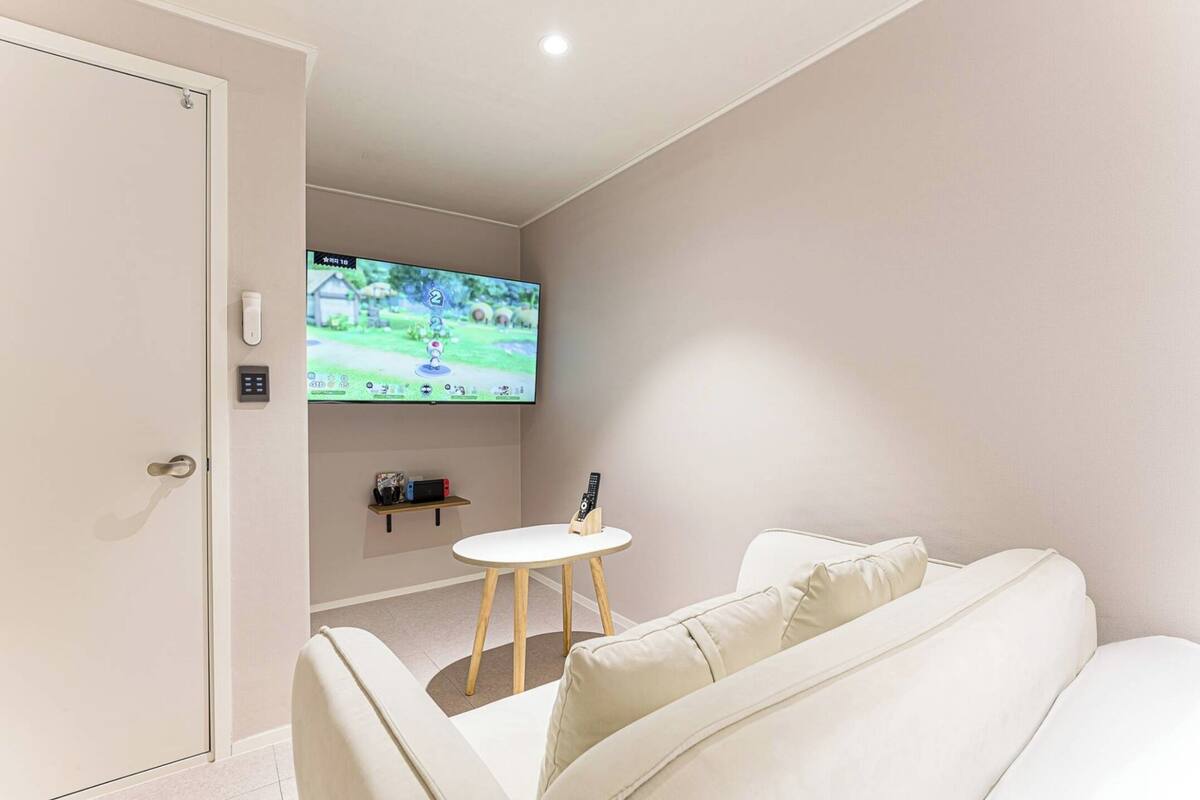 A modern living room with a white sofa, a small wooden table, and a mounted TV displaying a game.
