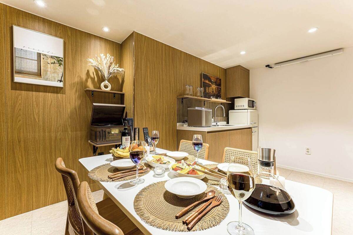 A dining area with a wooden wall, a kitchen in the background, and a table set for a meal.