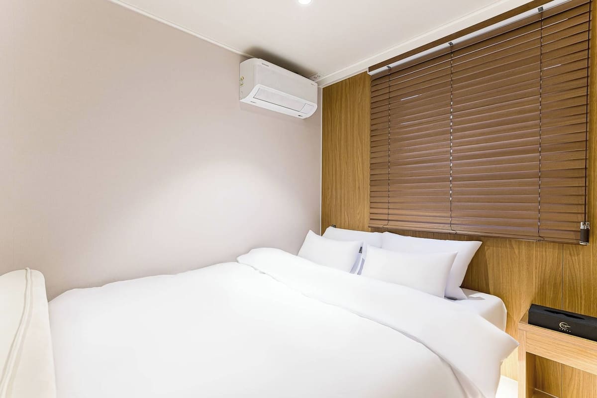 A hotel room with a bed, an air conditioning unit, wooden blinds, and a nightstand with a device.
